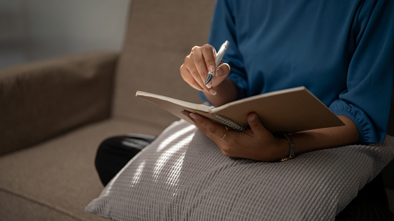 Close-up image of a woman in casual clothes is keeping her diary or taking notes on a notebook while sitting on a couch in her living room. Leisure concept