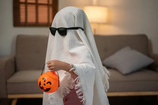 A baby little girl in a ghost costume with sunglasses is carrying a pumpkin bucket and walking around her living room. Spooky season, Halloween, trick or treat