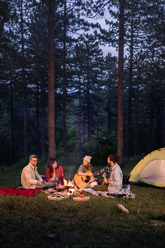 Young happy couples enjoying on camping during evening in the woods. Woman is playing a guitar. Copy space.