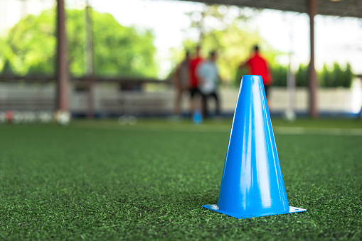 A blue obstracle cone which is placed on football turf ground, using for the movement and speed training session, photo with group of player as blur background. Sport practice activity scene.