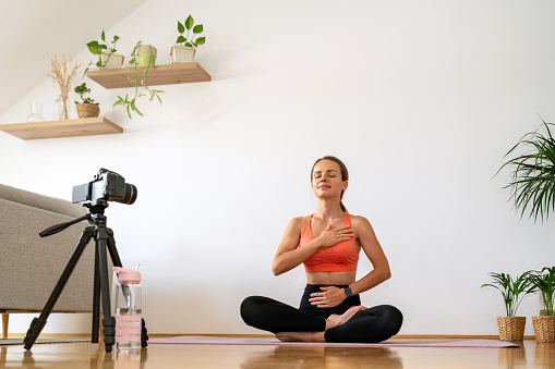 Young woman sits in lotus pose at home in front of video camera, meditating, doing breathing exercise.
