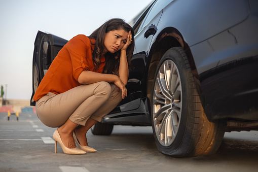 Portrait of a despair woman crouching next to her car with flat tire, problems on the road, deflated tire.