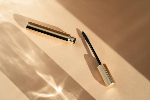 Mascara brush lies next to an open tube, on a beige background with shadows from sunlight. Copy space place for text cosmetic background