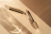 Mascara brush lies next to an open tube, on a beige background with shadows from sunlight. Copy space place for text cosmetic background.