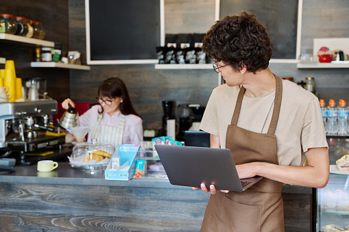 Young guy in an apron with laptop in his hands inside coffee shop cafeteria. Youth, work, online technology receiving orders delivery