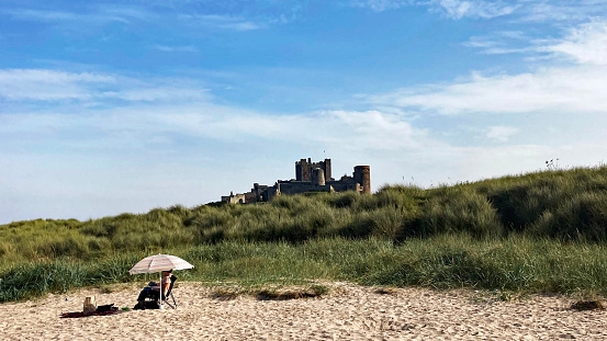 Bamburgh, Northumberland, England, September 8th. 2023: Solitary woman reading a book whilst sitting on the beach with Bamburgh Castle in the background