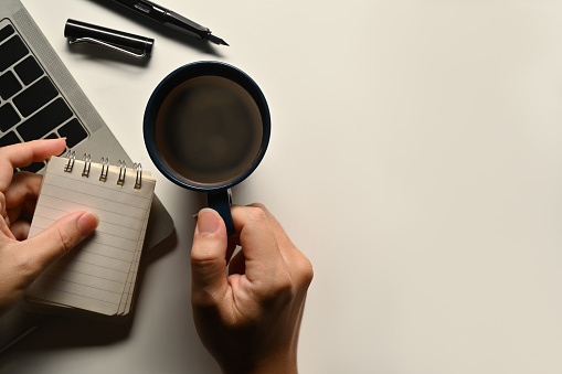 Above view of man hand holding cup of coffee and blank notepad over white office desk. Copy space for text.