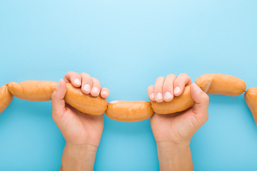 Young adult woman hands holding and showing string of raw fresh thick sausages for boiling. Light blue table background. Pastel color. Closeup. Point of view shot. Top down view.