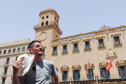 Mature man standing in front of city hall in Alicante, Spain. Cooling down with his hat.
