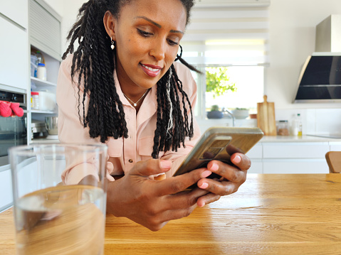 Shot of a happy Afro-American woman enjoying a glass of water while using a smartphone. The woman standing in the kitchen.