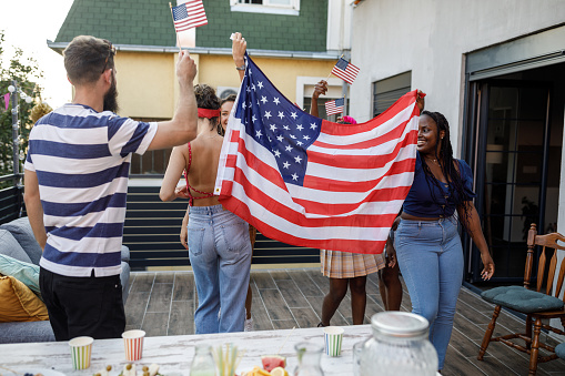 Candid shot of group of diverse, millennial people holding up and waving a big American flag, dancing and having fun while celebrating Independence Day on the balcony.