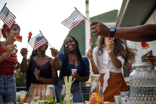 Candid shot of diverse group of joyful young people, friends, having fun, dancing and waving American flags while celebrating 4th of July on the balcony.