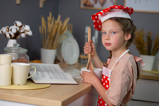 a girl in a red polka-dot apron and cap cooks in the kitchen. Nutrition and education of children. Children in the kitchen. Recipes for children. The child prepares food in the kitchen. Child protection. Children's Day.