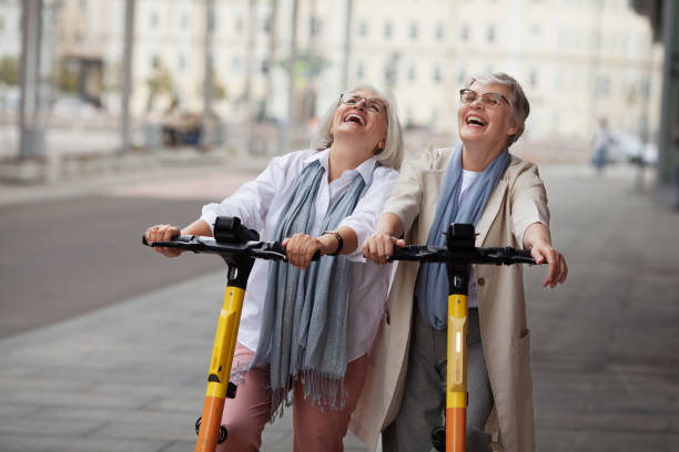 Portrait happy laughing Senior women with gray hair enjoying ride together on electric scooters outdoors. Two retired female friends at city street stock photo