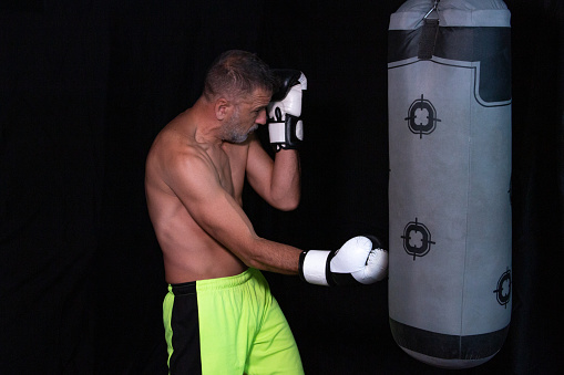 Mature man boxing, hitting a punching bag, shirtless on a black background