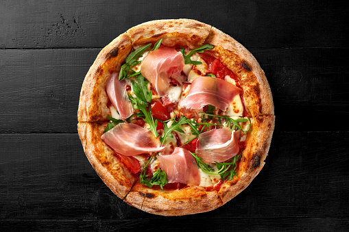 Delicious meat pizza with salami and thin slices of Italian prosciutto on base with pelati tomato sauce and mozzarella cheese garnished with fresh arugula leaves on black wooden background..