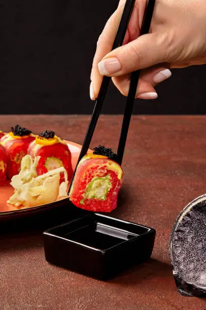 Photo of Female hand holding red rice sushi roll with tuna with chopsticks over soy sauce