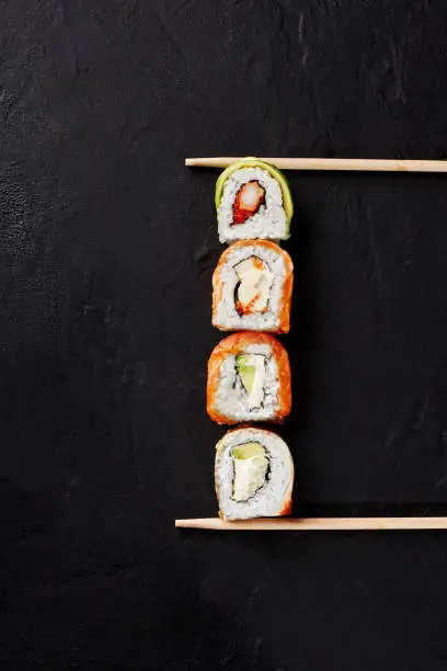 Photo of Four sushi rolls with salmon, eel, shrimp, avocado on black surface between chopsticks