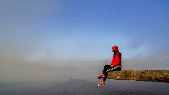 Hiker sitting on the edge of cliff at Phu Bak Dai, Loei, Thailand. A tourist woman sit alone at the cliff of mountain to see scenic view of sunset and range of mountain.