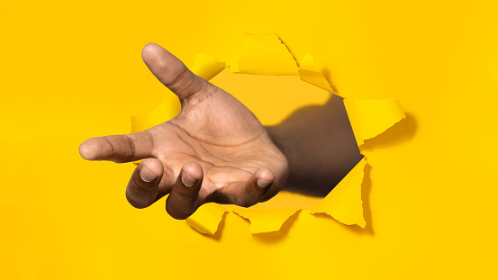 African american man stretching open palm through hole in torn yellow paper, offering helping hand, taking or giving something, asking for charity, reaching out for support
