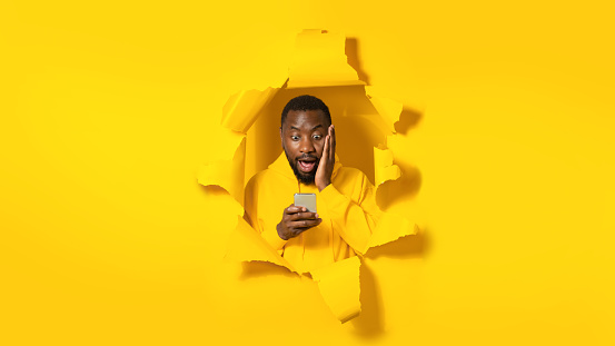 Surprised african american man standing in torn yellow paper hole, using cellphone and reading shocking news in social networks, panorama, copy space