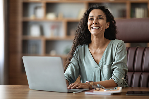 Emotional young lady in casual outfit sitting at worktable at office, typing on notebook keyboard and smiling, woman freelancer enjoying her remote job, copy space. Freelance concept