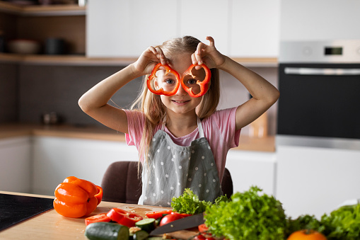 Cheerful european little kid puts pieces of pepper to her eyes, has fun in modern kitchen interior with organic vegetables. Cooking lesson, food blog, homemade lunch at home, health care and veganism