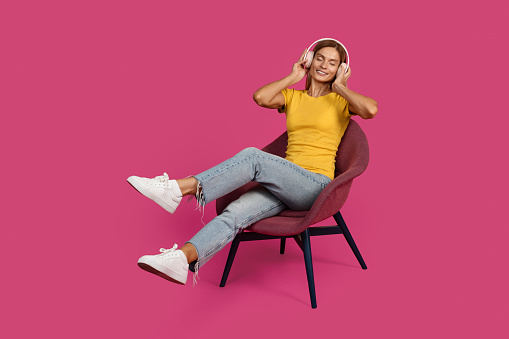 Happy adult caucasian blonde woman in casual and wireless headphones with closed eyes sits on armchair, listens music and enjoy free time, isolated on pink background, studio. Audio app for rest alone