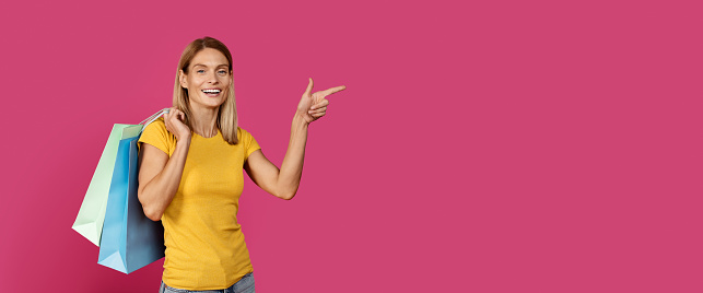 Glad mature caucasian blond female in yellow t-shirt with a lot of packages with purchases points finger at copy space, isolated on pink background, studio. Shopping, amazing offer, ad, huge sale