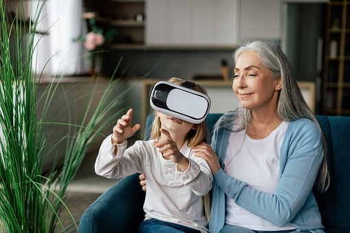 Happy caucasian small girl in vr glasses plays in virtual world with old grandma on sofa in living room interior. Rest and entertainment in free time, technology and online games at home due covid-19