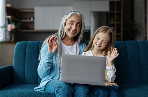 Happy european small girl and mature woman look at laptop, waving hand in living room interior. Meeting remotely, video call, relationship of granddaughter and grandma at home due covid-19 quarantine
