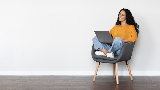 Cheerful pretty young woman freelancer using laptop at home, lady sitting in arm chair over white background, typing on keyboard and smiling, looking at copy space, panorama. Remote job concept