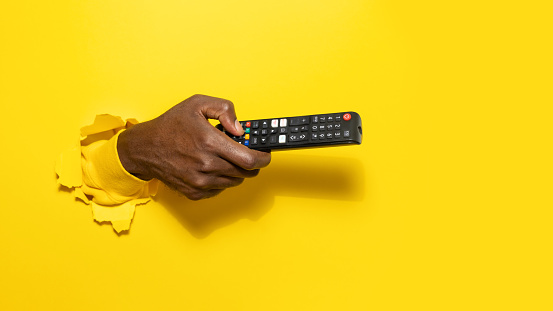 Black male hand holding remote control through the torn yellow paper background, pointing it aside at free space, space for your text or design, panorama