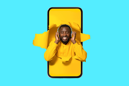 Joyful african american man in smartphone screen wearing wireless headphones and enjoying music with closed eyes over blue background. Music application concept