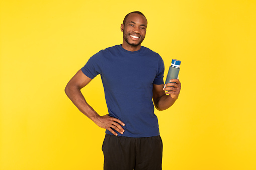 Fit African American Man Holding Water Bottle Posing Wearing Sportswear Standing Over Yellow Studio Background. Fitness Workout, Healthy Hydration Concept