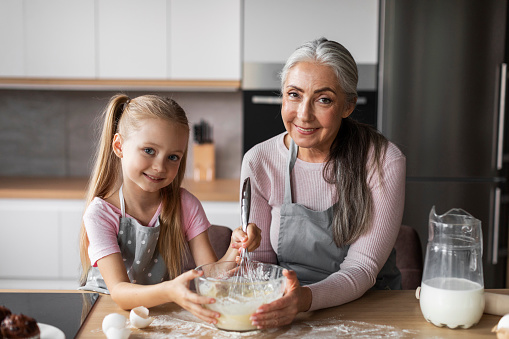 Cheerful pretty caucasian senior grandmother with small granddaughter make dough for baking, in modern kitchen interior. Chefs prepare sweets, girl and lady enjoy meal cooking lesson together at home