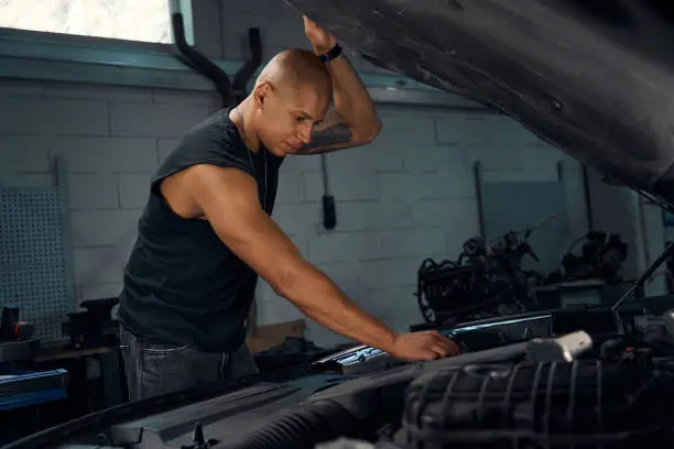 African American car-mechanic holding hood cover with one hand and checking terminals of battery with another, looking for break-down in client's car