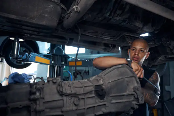 Concentrated African American car-mechanic resting standing under car and leaning on some detail, engaged in automobiles maintenance and repairing, motor vehicle service station