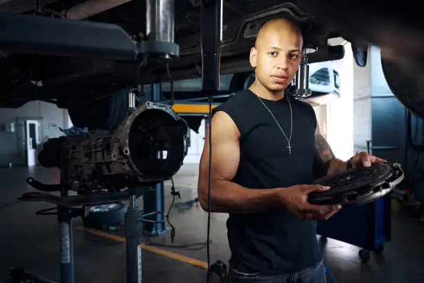 Concerned Africa American car-mechanic removed clutch disc from car and examining on breakage presence, seriously looking at camera finding problem, motor vehicle service station