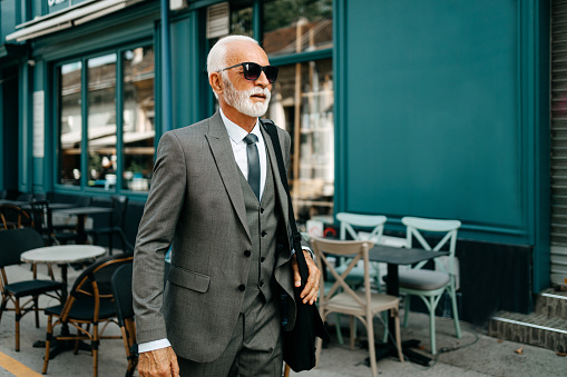 Senior Caucasian businessman wearing sunglasses and a modern suit is walking through a city street, carrying a laptop bag.