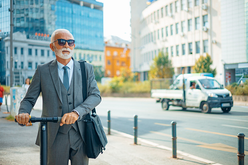 Senior Caucasian businessman wearing sunglasses and a modern suit is going through the city on an electric scooter.