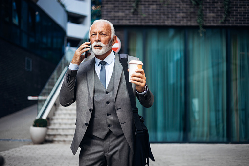 Senior Caucasian businessman walking through the city and talking on the phone with a serious look on his face. He's holding a coffee cup.