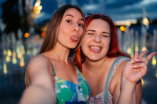 Portrait of two beautiful female friends taking selfies with their tongues out using smart phone in front of a city fountain at dusk.