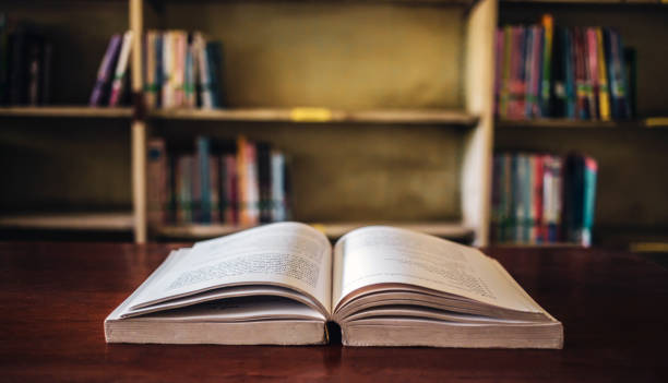 istock On a wooden table, there's an open book with a library visible in the background. A stack of books nearby adds to the theme of educational learning. 1703952356