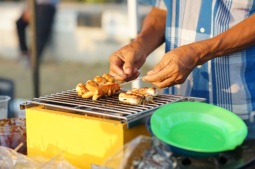 Asian male street food seller grilling sausages using a sausage grill machine at his stall