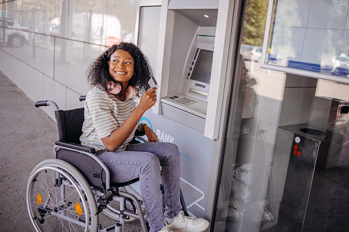 Young black woman in wheelchair picking up money from ATM machine