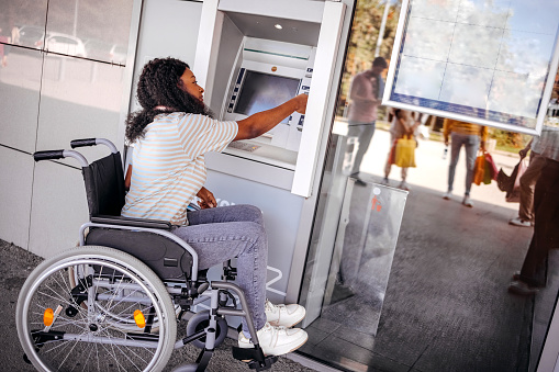 Young black woman in wheelchair picking up money from ATM machine