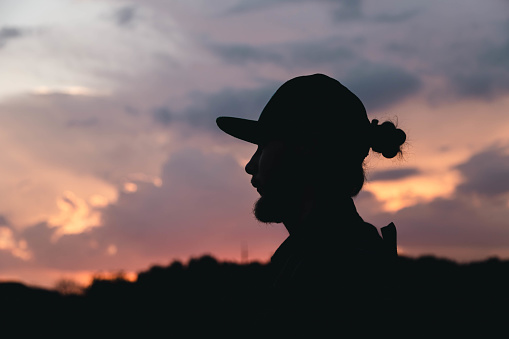 A bearded man with a bun and a cap at sunset on the sky background, a black silhouette of a man.