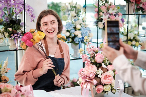 E-commerce business, one young White female florist demonstrates and shows floral arrangements via online live streaming with smartphone application in bright flower shop, beautiful blossoms store.
