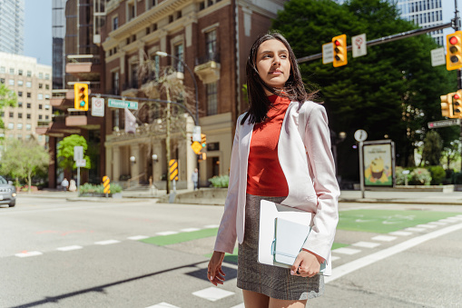 Confident Indian student girl wearing stylish clothes, holding laptop computer looking away standing on the street, copy space. Successful business and education